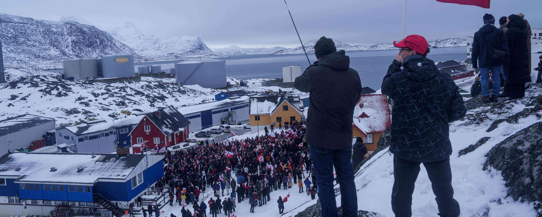 Zwei Menschen wehen auf einem Hügel grönländische Fahnen, im Hintergrund ist unter ihnen eine Menge von Demonstranten zu sehen.