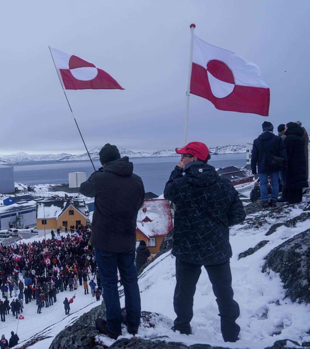 Zwei Menschen wehen auf einem Hügel grönländische Fahnen, im Hintergrund ist unter ihnen eine Menge von Demonstranten zu sehen.