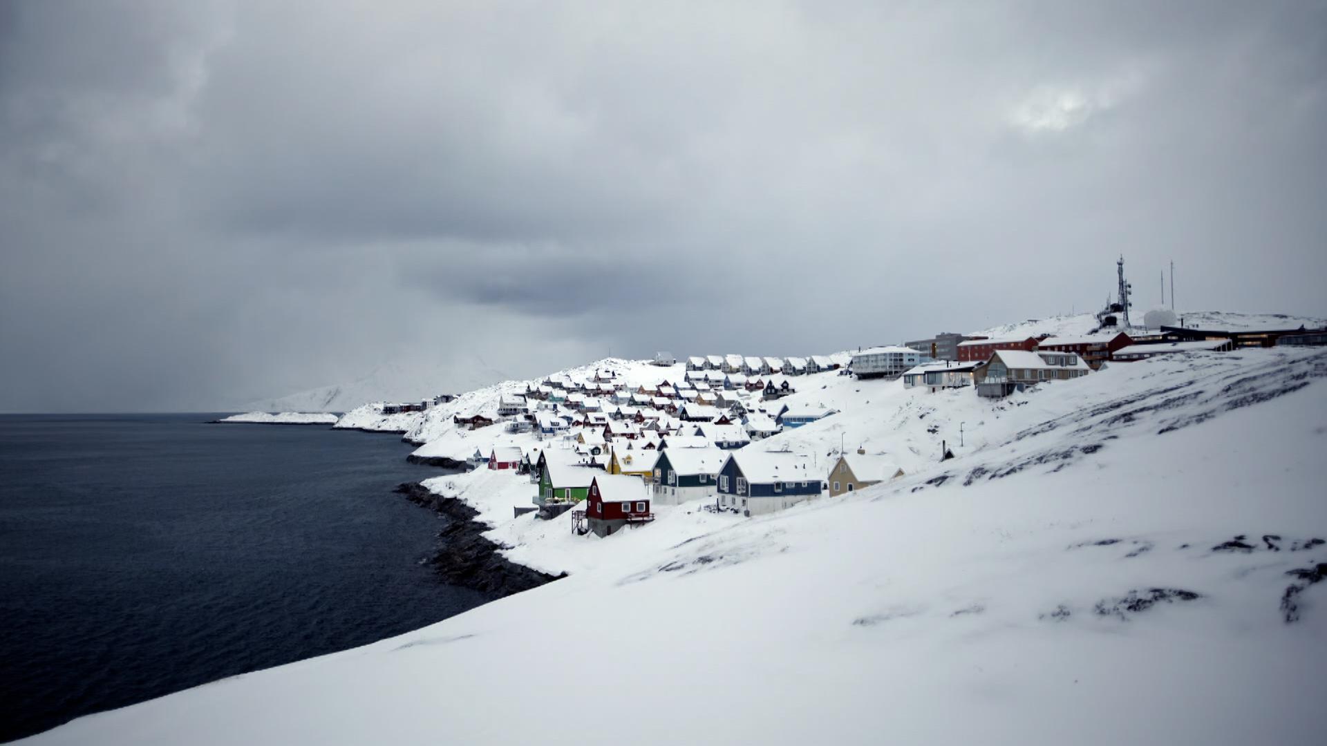 Zu sehen ist eine verschneite Küstenregion in Grönland: links das Meer, rechts Häuser.