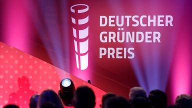 Das Veranstaltungsvisual des Deutschen Gründerpreises ist in kräftigen Rot-, Pink- und Violetttönen gestaltet. Im Hintergrund sind angeschnittene weiße Logos und Schriftzüge des Preises zu sehen. Im unscharfen Vordergrund befinden sich die Köpfe eines Publikums sowie eine auf die Bühne gerichtete Kamera und ein heller Scheinwerfer. Der Deutsche Gründerpreis wird am 8. September 2026 im ZDF-Hauptstadtstudio in Berlin verliehen.