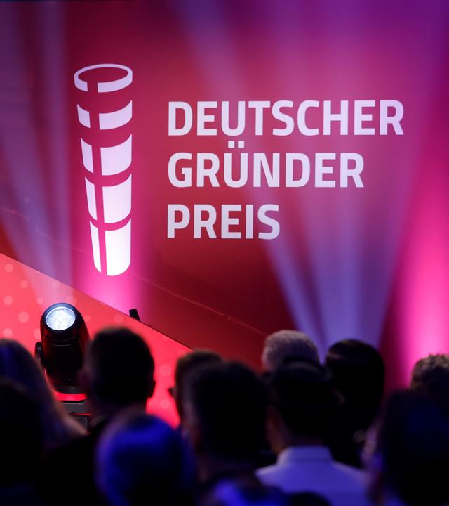 Das Veranstaltungsvisual des Deutschen Gründerpreises ist in kräftigen Rot-, Pink- und Violetttönen gestaltet. Im Hintergrund sind angeschnittene weiße Logos und Schriftzüge des Preises zu sehen. Im unscharfen Vordergrund befinden sich die Köpfe eines Publikums sowie eine auf die Bühne gerichtete Kamera und ein heller Scheinwerfer. Der Deutsche Gründerpreis wird am 8. September 2026 im ZDF-Hauptstadtstudio in Berlin verliehen.