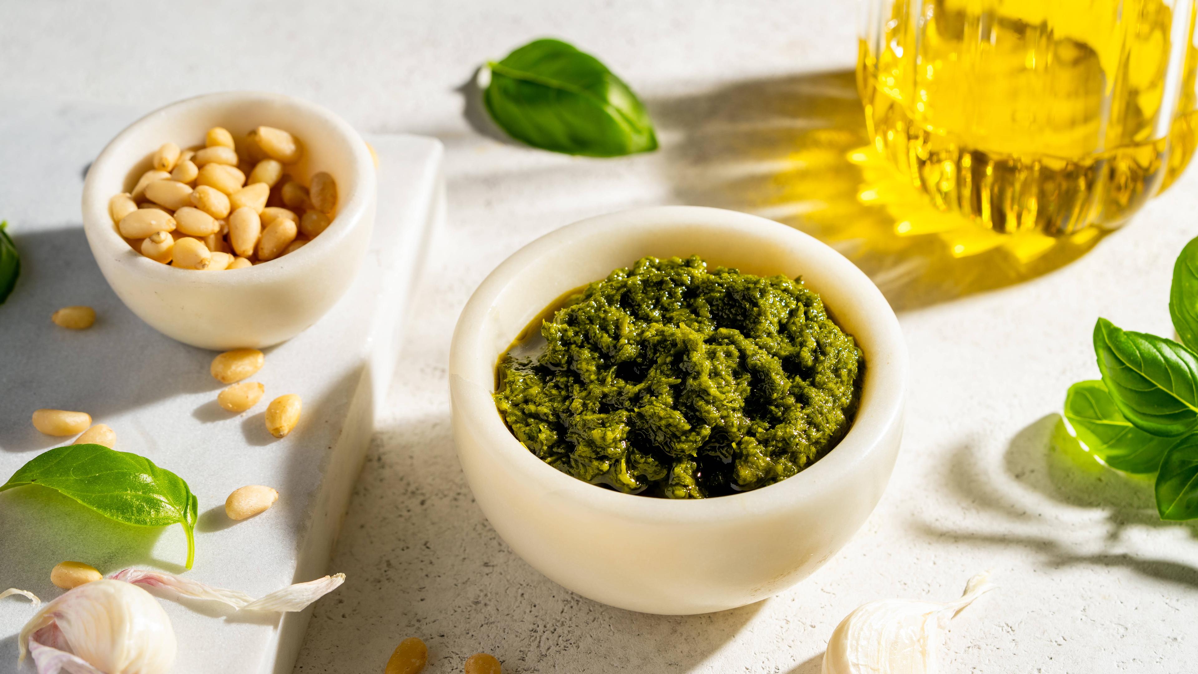 Eine Schüssel mit grünem Pesto, eine Schüssel mit Pinienkernen, Basilikumblättern, Knoblauch und eine Glasflasche mit Öl von oben.
