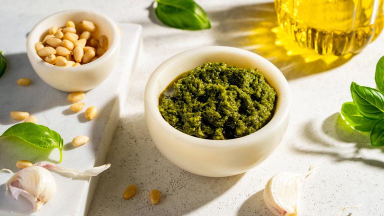 Eine Schüssel mit grünem Pesto, eine Schüssel mit Pinienkernen, Basilikumblättern, Knoblauch und eine Glasflasche mit Öl von oben.