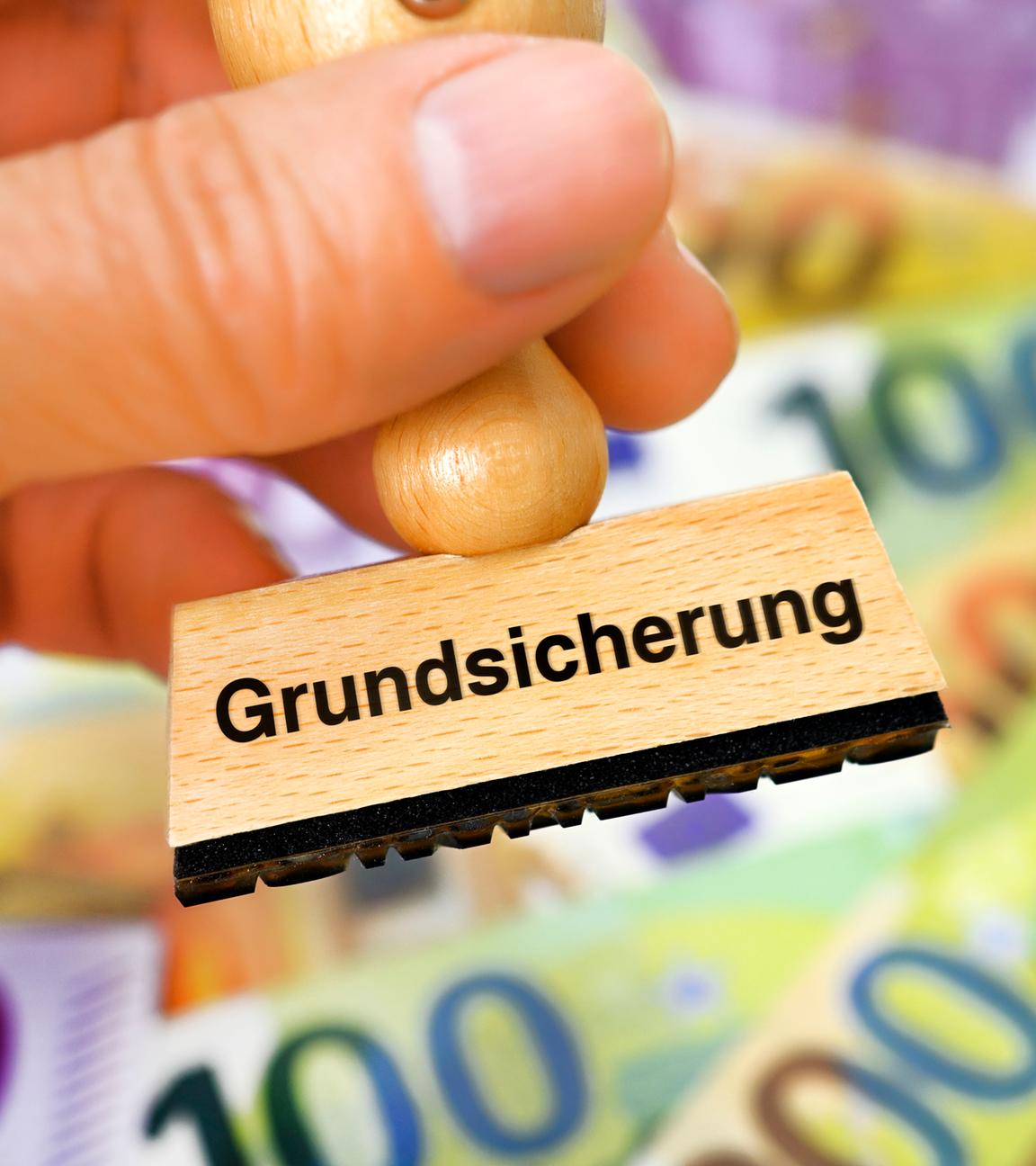 Symbolbild: Hand hält Stempel mit Aufschrift Grundsicherung vor Geldscheinen