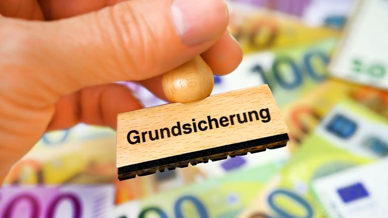 Symbolbild: Hand hält Stempel mit Aufschrift Grundsicherung vor Geldscheinen