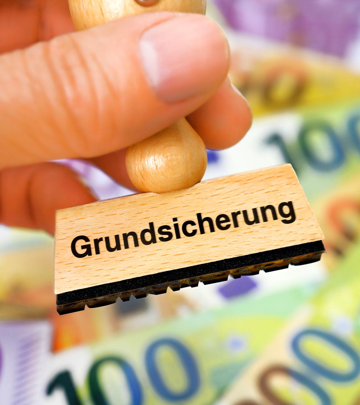 Eine Hand hält Stempel mit Aufschrift "Grundsicherung" vor einigen Geldscheinen.