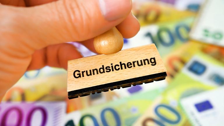 Eine Hand hält Stempel mit Aufschrift "Grundsicherung" vor einigen Geldscheinen.