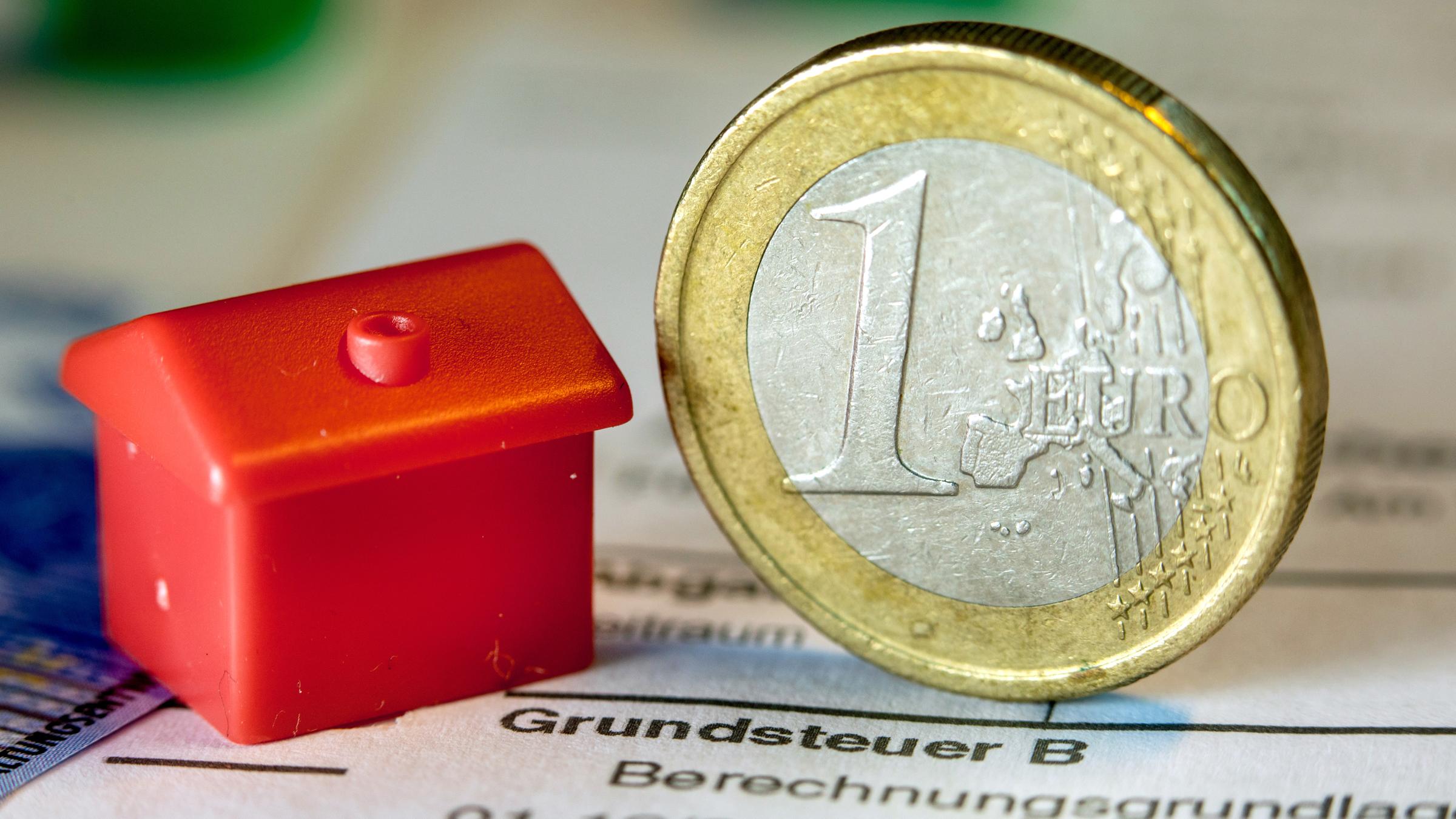 Eine Euromünze und ein kleines Spielzeughaus liegen auf einem Dokument zur Grundsteuer.