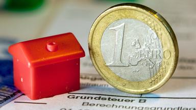 Eine Euromünze und ein kleines Spielzeughaus liegen auf einem Dokument zur Grundsteuer.