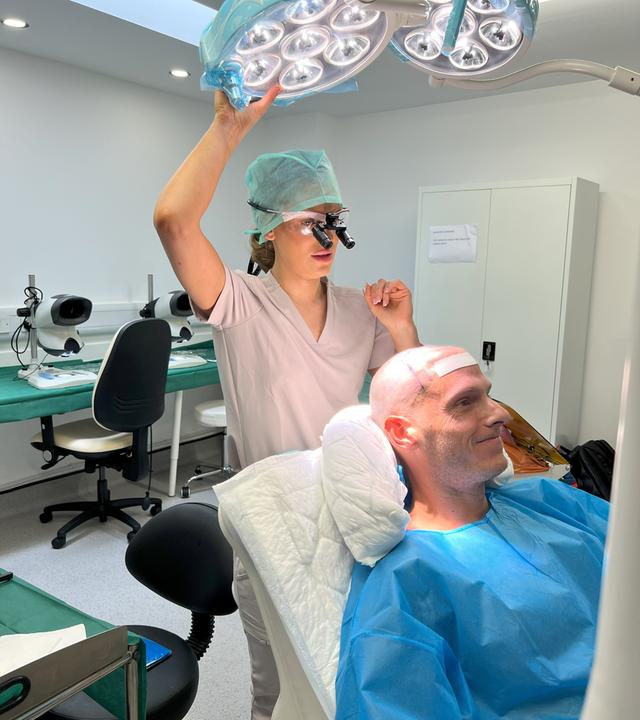 Ein Mann mit Glatze liegt auf einer Liege in einer Klinik. Hinter ihm steht eine Klinikmitarbeiterin und greift nach einer großen, über der Liege angebrachten Leuchte. Sie trägt eine Haube und eine chirurgische Brille.