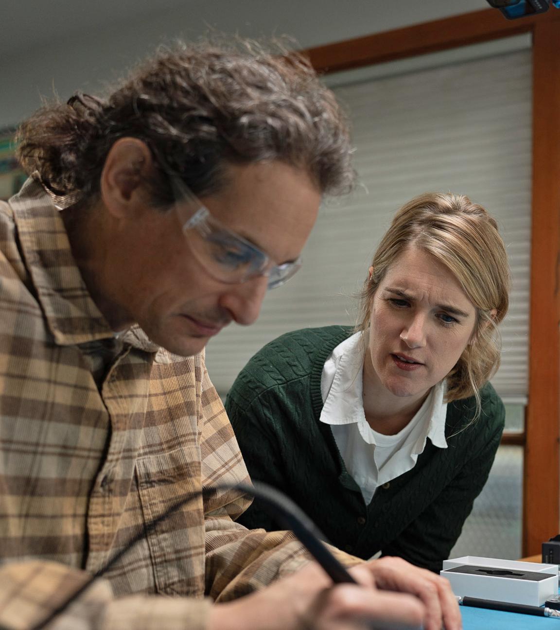 Joe Grand mit Schutzbrille und kariertem Hemd. Er arbeitet an einem Werktisch mit einem Lötgerät in der Hand. Neben ihm nach vorn gebeugt eine Frau, die interessiert seine Arbeit verfolgt.