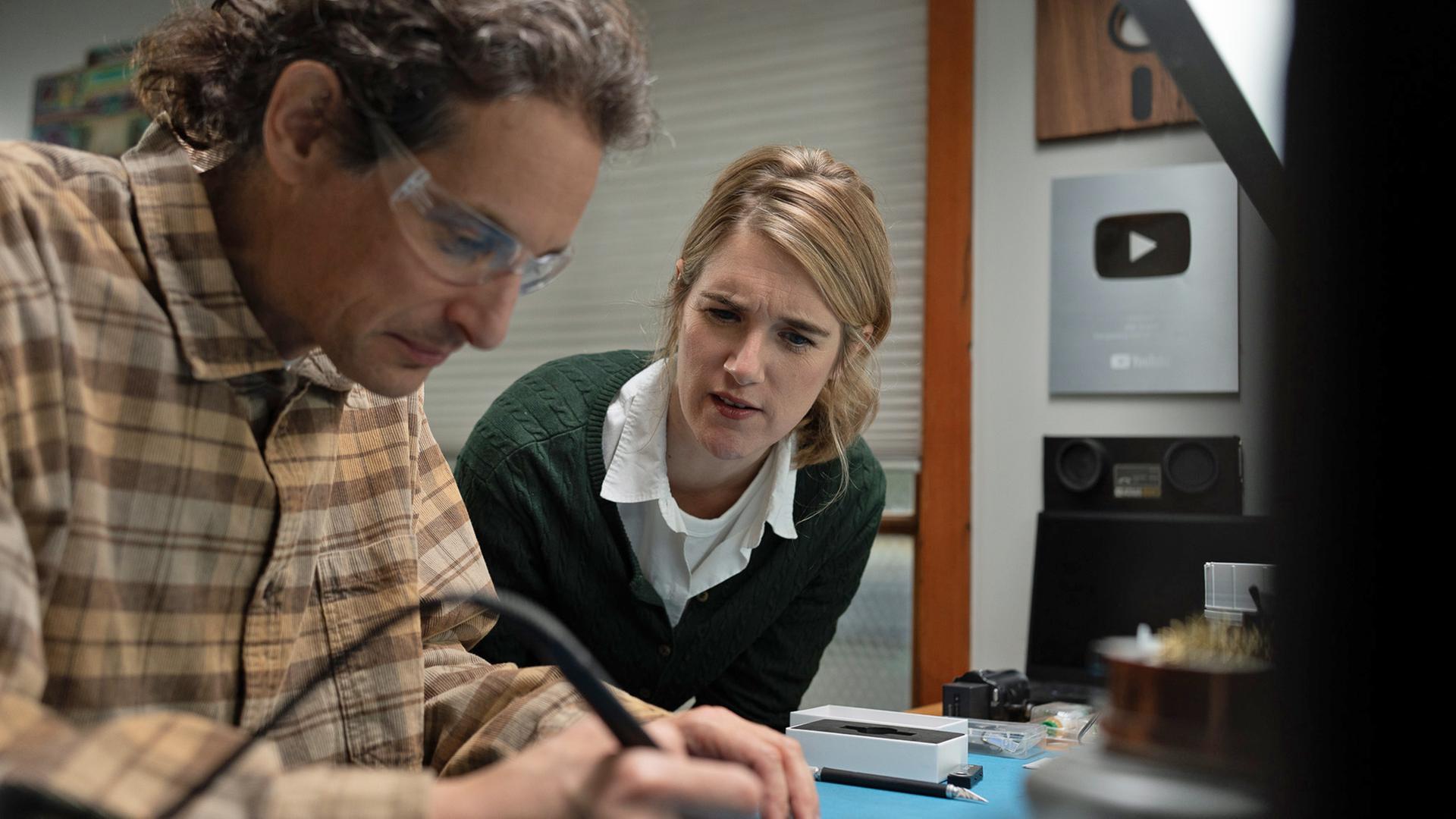 Joe Grand mit Schutzbrille und kariertem Hemd. Er arbeitet an einem Werktisch mit einem Lötgerät in der Hand. Neben ihm nach vorn gebeugt eine Frau, die interessiert seine Arbeit verfolgt.