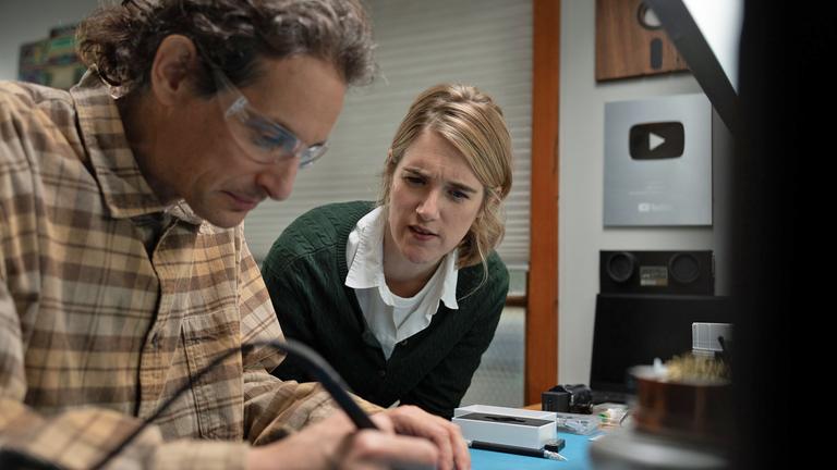 Joe Grand mit Schutzbrille und kariertem Hemd. Er arbeitet an einem Werktisch mit einem Lötgerät in der Hand. Neben ihm nach vorn gebeugt eine Frau, die interessiert seine Arbeit verfolgt.