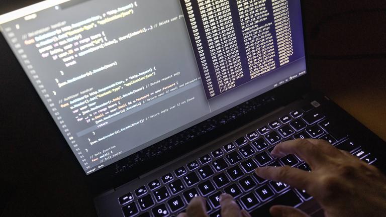 Symbolbild: Quellcode eines Webservers sowie die Windows-Eingabeaufforderung sind einem Laptop im Dunkeln zu sehen., 