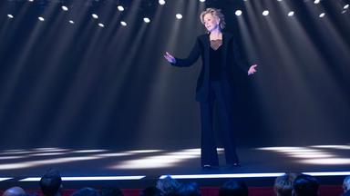 Ausschnitt aus der Dramedy-Serie "Hacks" - Deborah (Jean Smart) steht in einem eleganten schwarzen Outfit mit leicht besorgtem Gesichtsausdruck und weit geöffneten Armen auf einer Bühne, die von zahlreichen Scheinwerfern im Hintergrund erhellt wird.