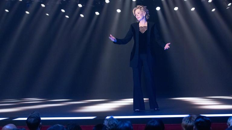 Ausschnitt aus der Dramedy-Serie "Hacks" - Deborah (Jean Smart) steht in einem eleganten schwarzen Outfit mit leicht besorgtem Gesichtsausdruck und weit geöffneten Armen auf einer Bühne, die von zahlreichen Scheinwerfern im Hintergrund erhellt wird.