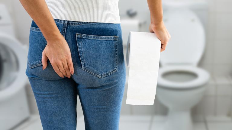 Eine Frau wird von hinten gezeigt. Sie greift sich über die Hose an den Po. Sie hält eine Rolle Klopapier in der Hand und schaut in Richtung einer Toilette.