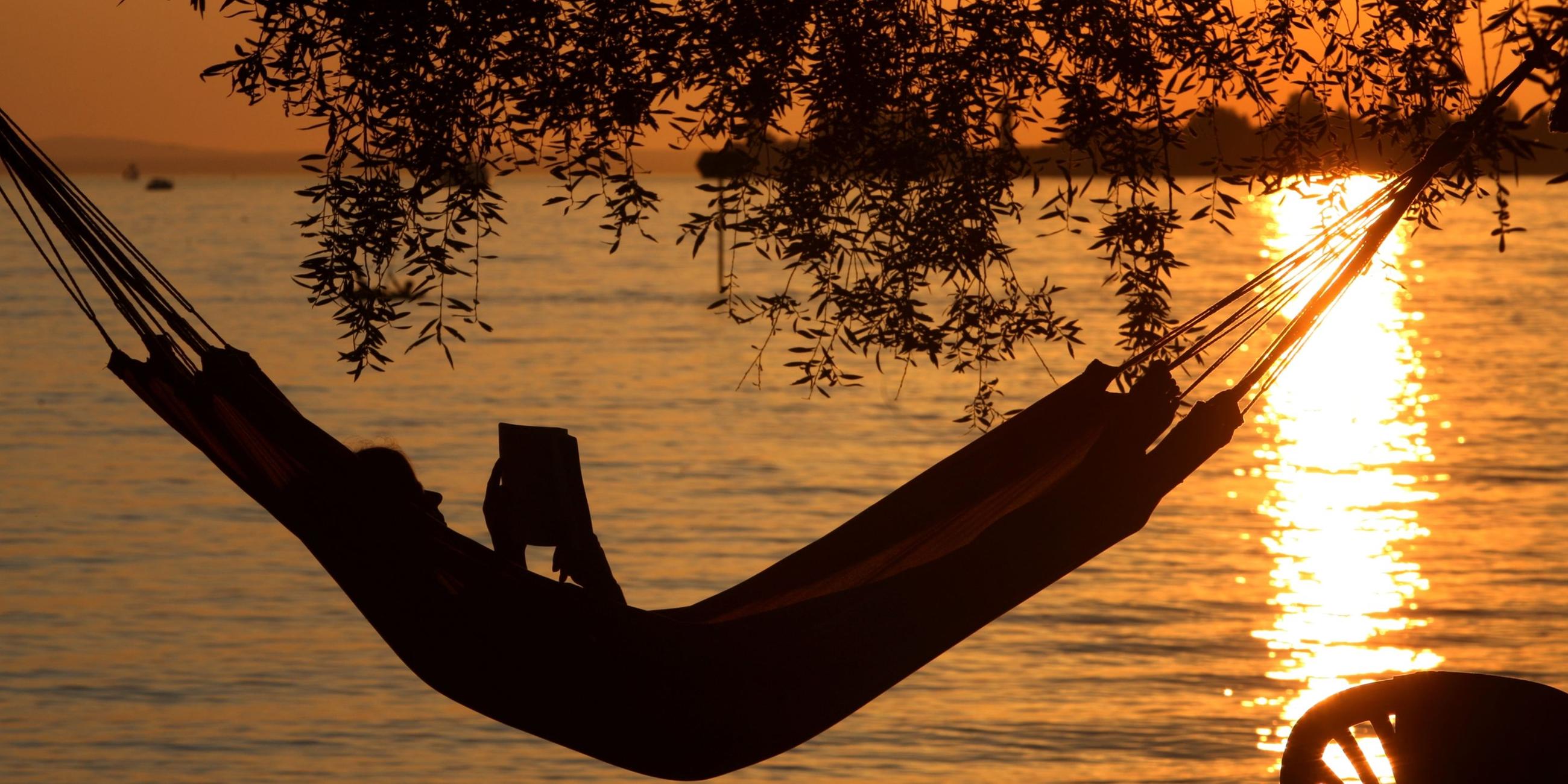 Eine Frau liest in einer Hängematte am Badestrand eines Campingplatzes ein Buch. 