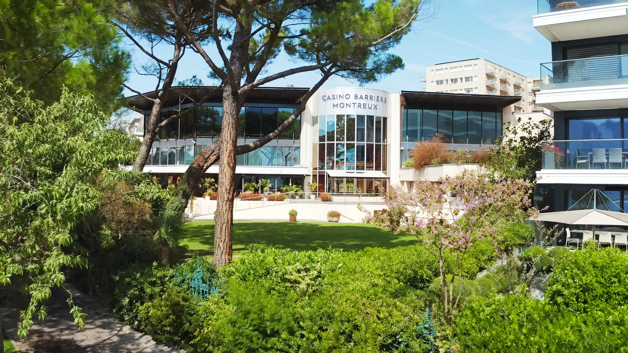 Die Fassade des Casinoss Barrière Montreux in der Schweiz mit großen Fenstern und geraden Architekturlinien. Davor grüne Bäume und Büsche.