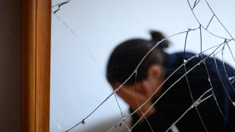  Eine Frau sitzt neben einem gesprungenen Spiegel auf einem Bett und hält ihren Kopf in den Händen (gestellte Szene).