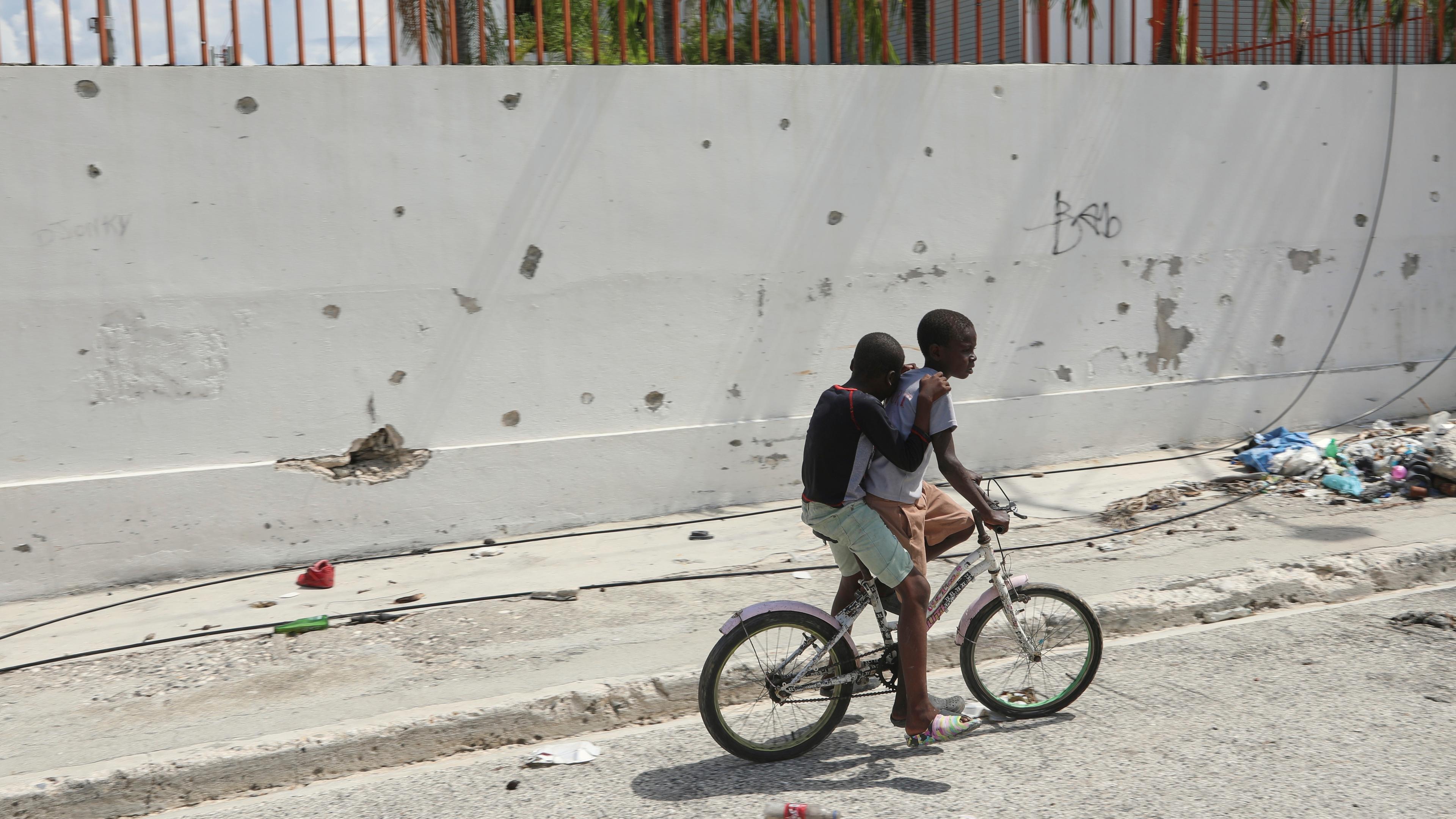 In Haiti fahren zwei Kinder auf einem Fahrrad an einer Wand vorbei, die von Schusslöchern durchlöchert ist.