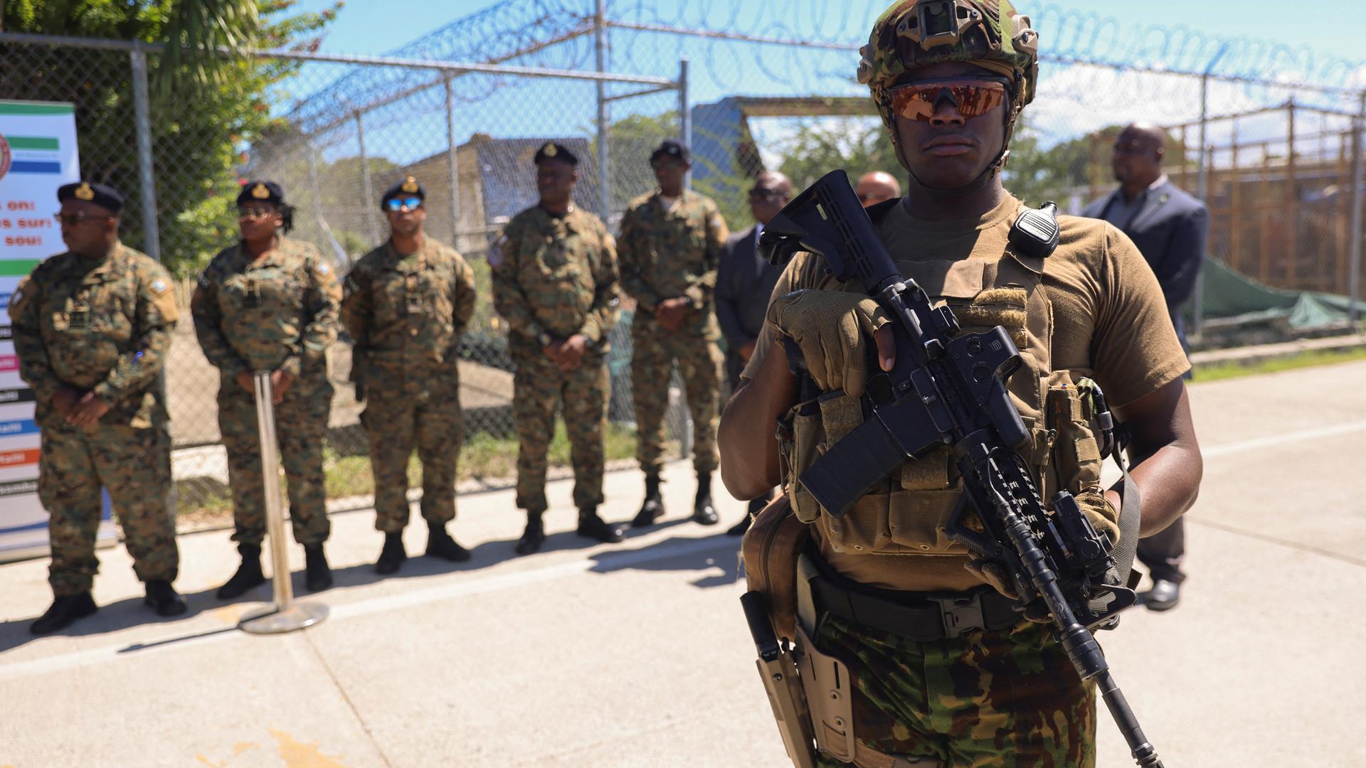 Schwer bewaffnete Männer stehen vor einem Maschendrahtzaun in Tarnkleidung.