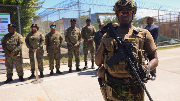 Schwer bewaffnete Männer stehen vor einem Maschendrahtzaun in Tarnkleidung.