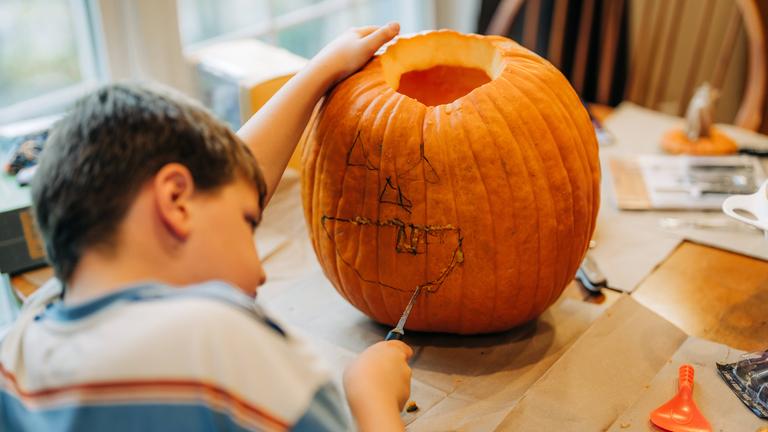 Ein Kind sitzt am Esstisch und schnitzt eine Fratze in einen Kürbis.