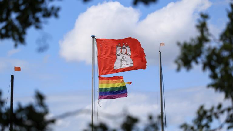 Eine Regenbogenflagge und die Hamburger Flagge hängen an einem Fahnenmast.