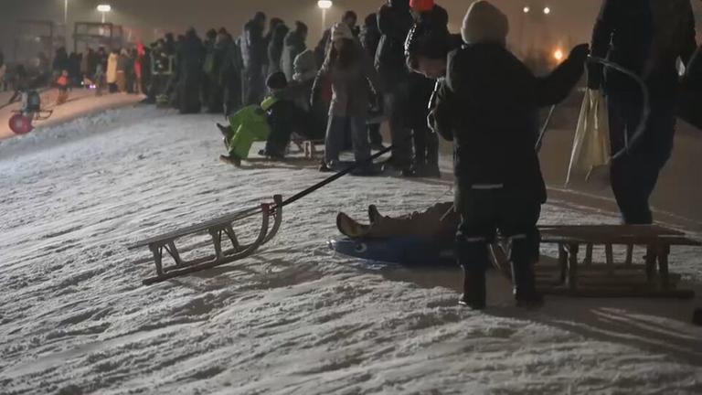 Menschen stehen mit Schlitten auf einem verschneiten Hügel. Der Hügel wird mit Scheinwerfern beleuchtet.