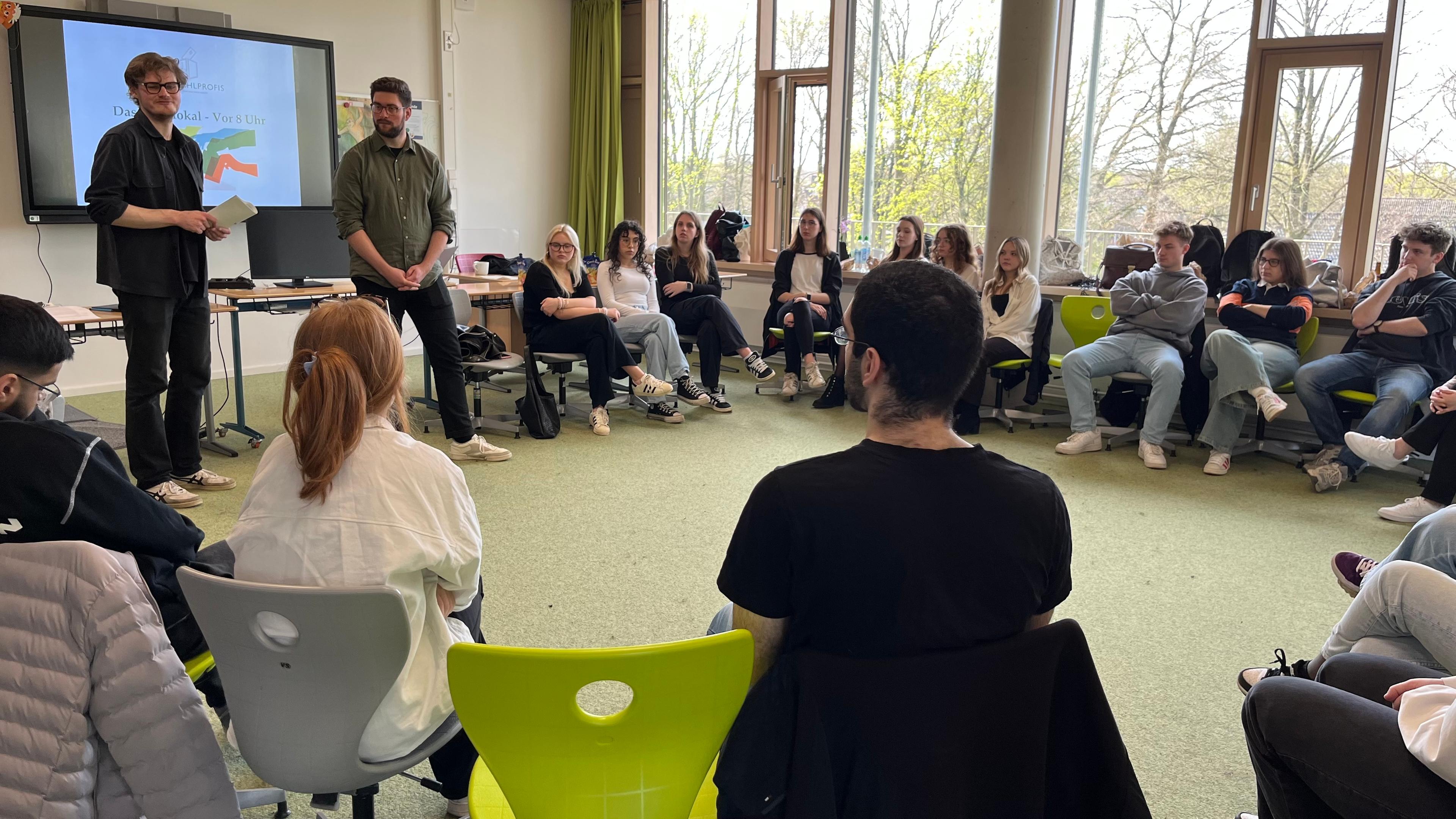 Eine Schulklasse sitzt im Stuhlkreis im Klassenzimmer. Zwei Männer halten einen Vortrag.