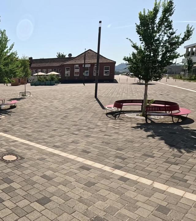 Marga-von-Etzdorf-Platz in Stuttgart Bad-Canntstatt: Eine große, weitgehend leere Fläche mit dunklem Backsteinboden, auf der nur drei kleine Bäume mit Sitzbank stehen, als ob das reichen würde, damit sich der Platz nicht im Sommer stark erhitzt.