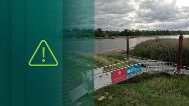 Ein Anlegesteg aus Metall an einem Flussufer, links und rechts von hohen Gräsern umgeben.