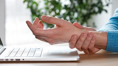 Eine Person sitzt vor dem Laptop und hält sich das Handgelenk.