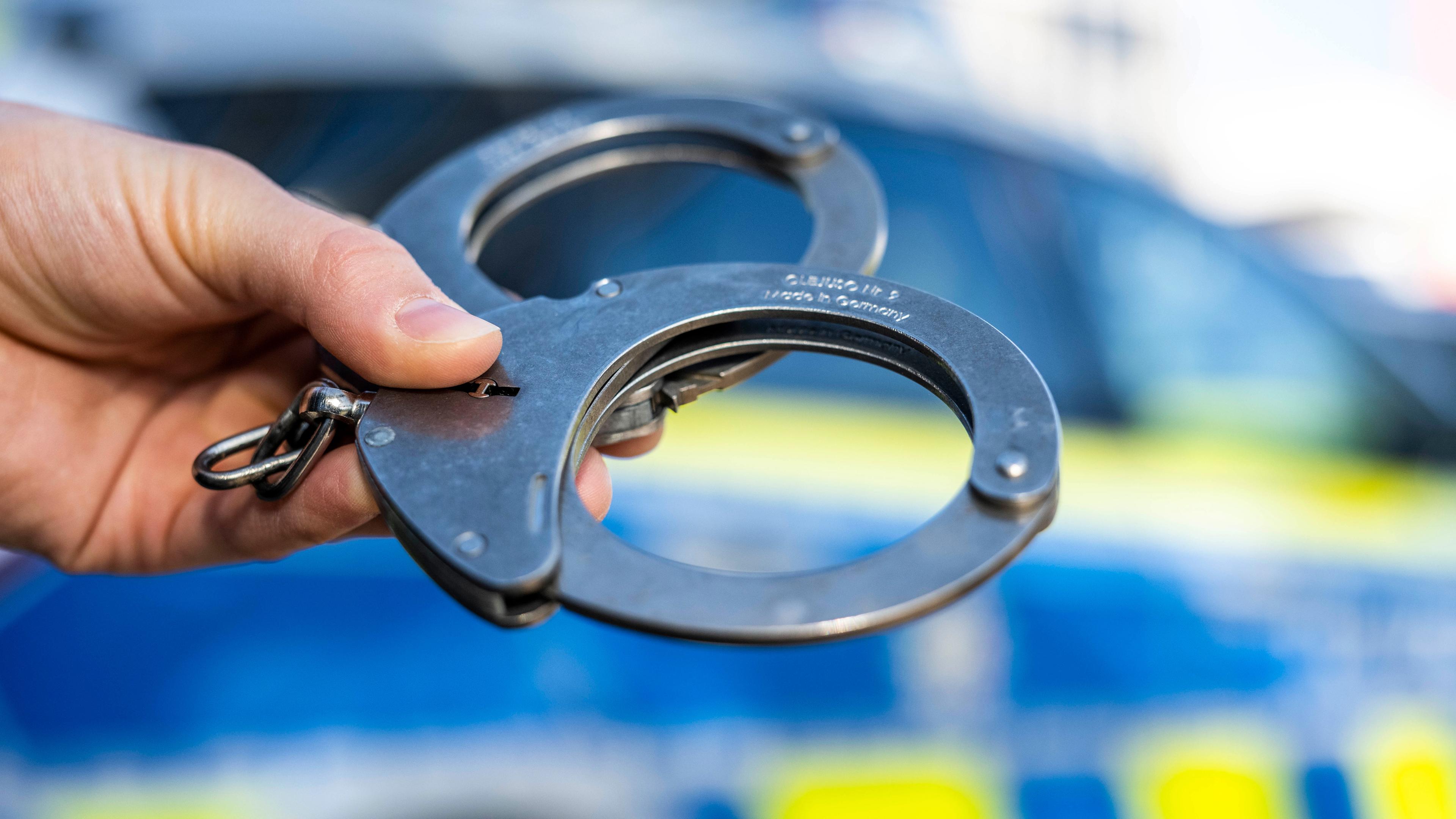 Hand hält Handschellen vor einen Streifenwagen