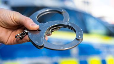 Hand hält Handschellen vor einen Streifenwagen
