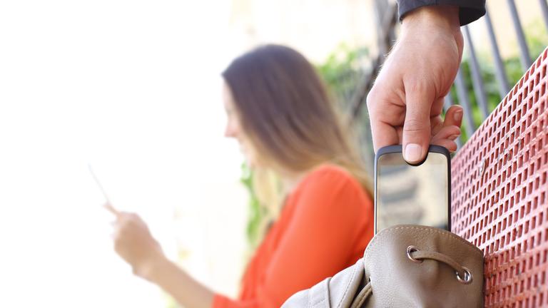 Ein Mensch klaut ein Handy aus der Tasche einer Frau auf einer Parkbank.
