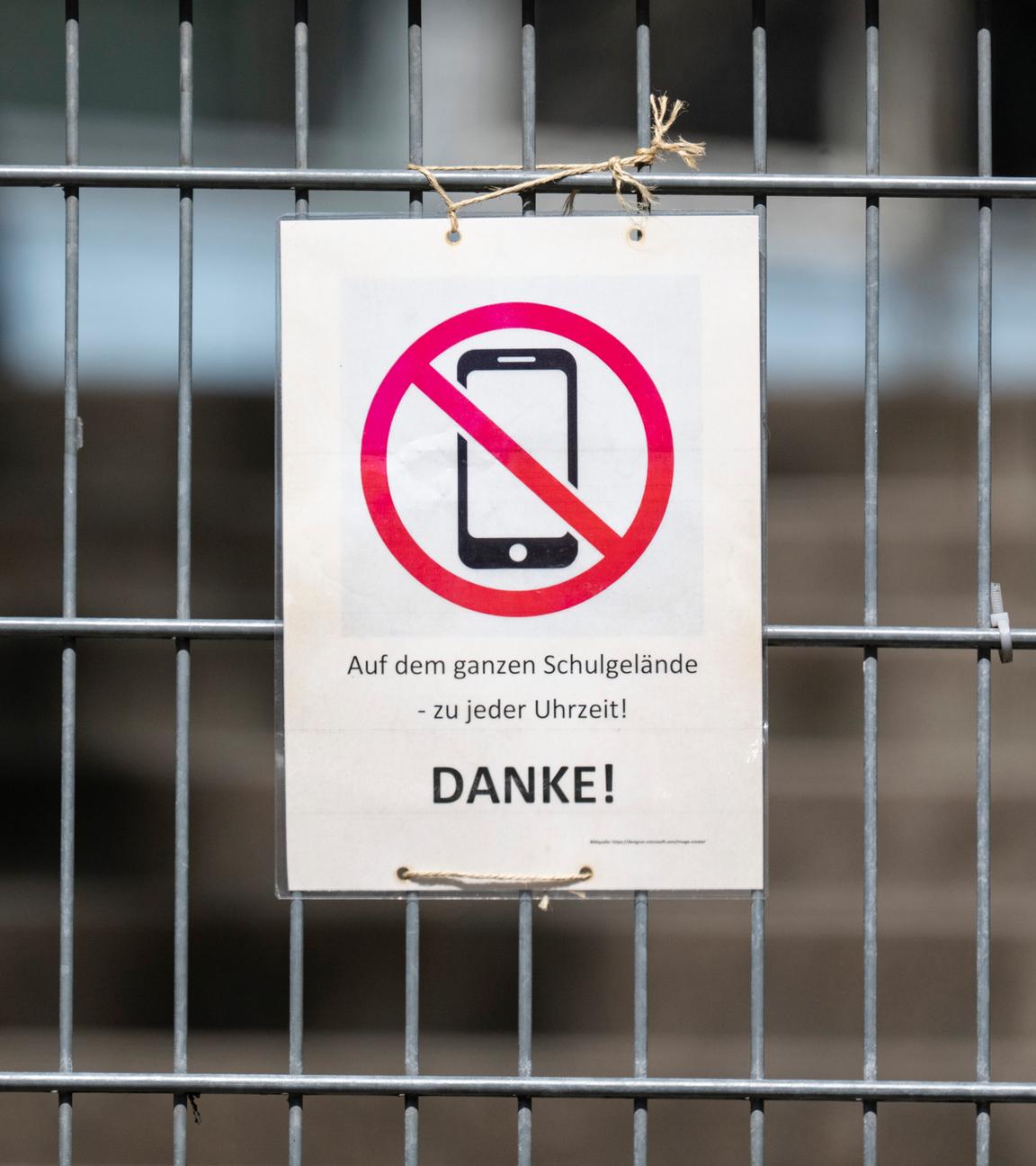 08.04.2025, Baden-Württemberg, Stuttgart: Ein Schild mit einem durchgestrichenen Smartphone und der Aufschrift "Auf dem ganzen Schulgelände - zu jeder Uhrzeit! DANKE!" hängt am Zugangstor zum Schulhof eines Gymnasiums und weist auf ein generelles Handyverbot hin.
