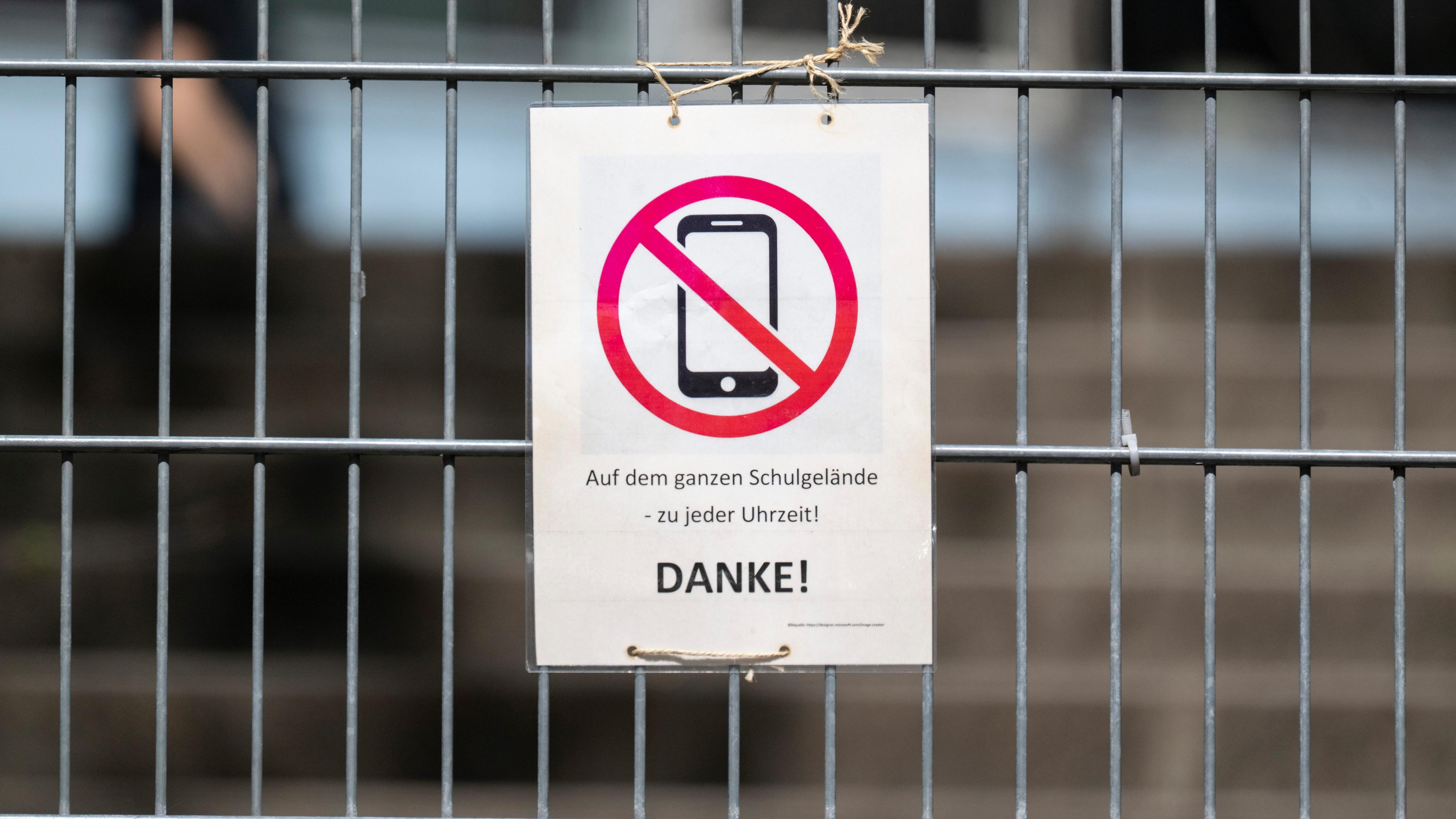 08.04.2025, Baden-Württemberg, Stuttgart: Ein Schild mit einem durchgestrichenen Smartphone und der Aufschrift "Auf dem ganzen Schulgelände - zu jeder Uhrzeit! DANKE!" hängt am Zugangstor zum Schulhof eines Gymnasiums und weist auf ein generelles Handyverbot hin.
