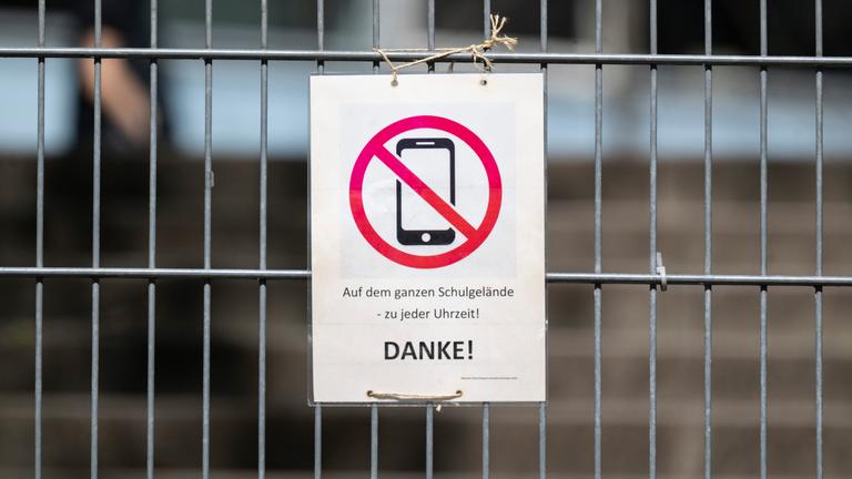 08.04.2025, Baden-Württemberg, Stuttgart: Ein Schild mit einem durchgestrichenen Smartphone und der Aufschrift "Auf dem ganzen Schulgelände - zu jeder Uhrzeit! DANKE!" hängt am Zugangstor zum Schulhof eines Gymnasiums und weist auf ein generelles Handyverbot hin.