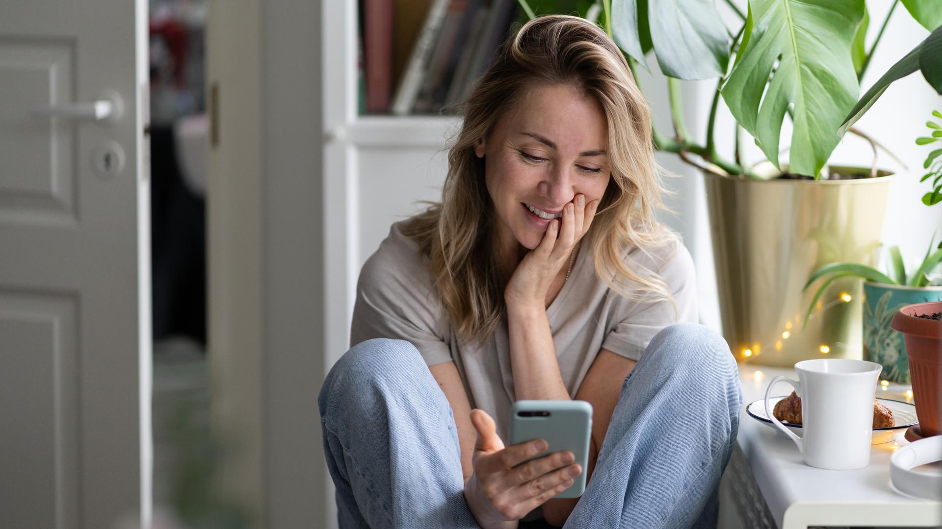 Eine Frau sitzt lächelnd in einem Zimmer und schaut auf ihr Handy.