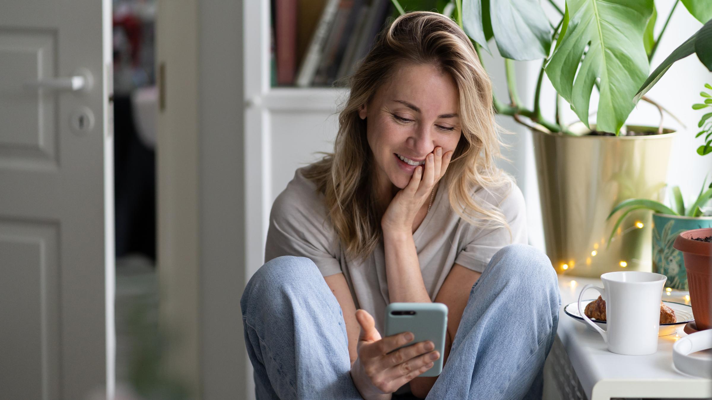Eine Frau sitzt lächelnd in einem Zimmer und schaut auf ihr Handy.