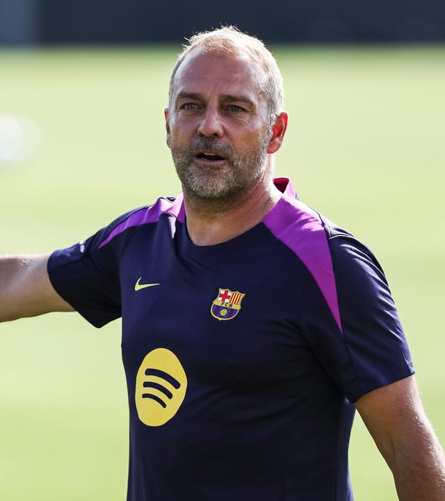 Der Trainer von FC Barcelona, Hansi Flick steht in einem Barcelona Trikot auf einem Fußballplatz.