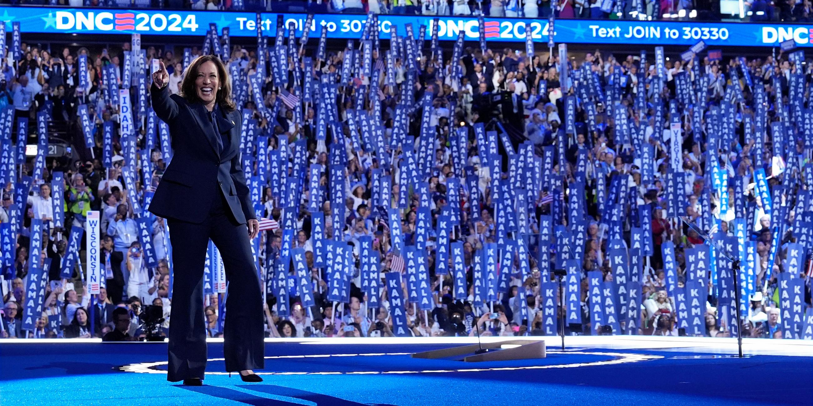 Kamala Harris steht in einem dunklen Anzug auf der Bühne und winkt in die Menge. 