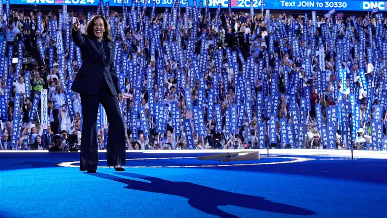 Kamala Harris steht in einem dunklen Anzug auf der Bühne und winkt in die Menge. 