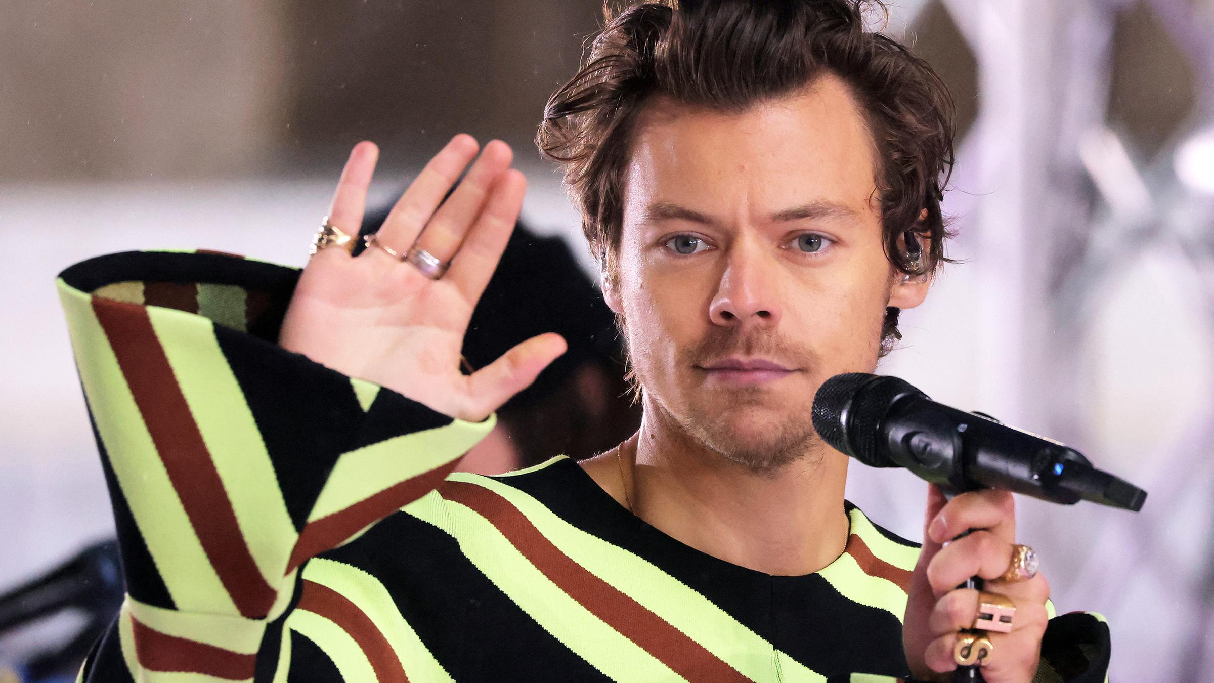 Harry Styles hinter einem Mikrofon, er winkt mit einer Hand.