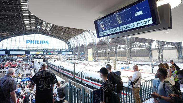 Hamburg: Bahnreisende stehen an den Bahnsteigen im Hamburger Hauptbahnhof, während auf einer Anzeigetafel auf den beeinträchtigten Bahnverkehr  hingewiesen wird