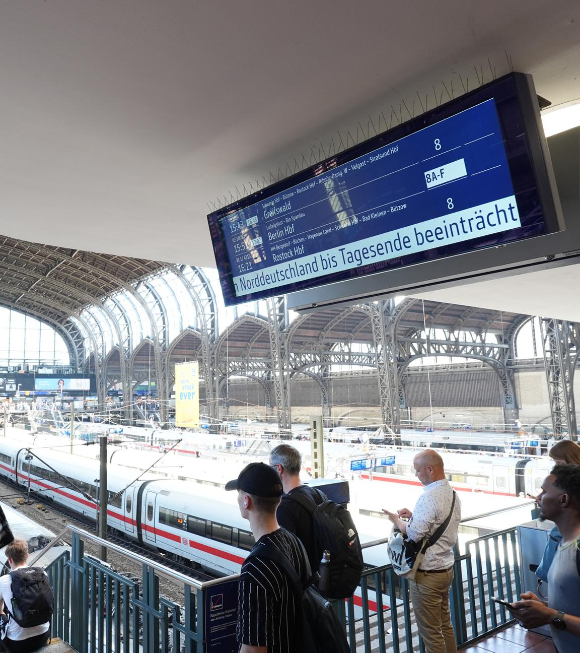Hamburg: Bahnreisende stehen an den Bahnsteigen im Hamburger Hauptbahnhof, während auf einer Anzeigetafel auf den beeinträchtigten Bahnverkehr  hingewiesen wird