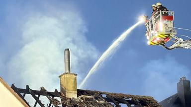 Einsatzkräfte der Feuerwehr bekämpfen einen Brand an einem Wohnhaus. Von einer Drehleiter aus wird Wasser auf den teilweise zerstörten Dachstuhl gespritzt, während Rauch aufsteigt.