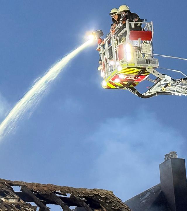 Einsatzkräfte der Feuerwehr bekämpfen einen Brand an einem Wohnhaus. Von einer Drehleiter aus wird Wasser auf den teilweise zerstörten Dachstuhl gespritzt, während Rauch aufsteigt.